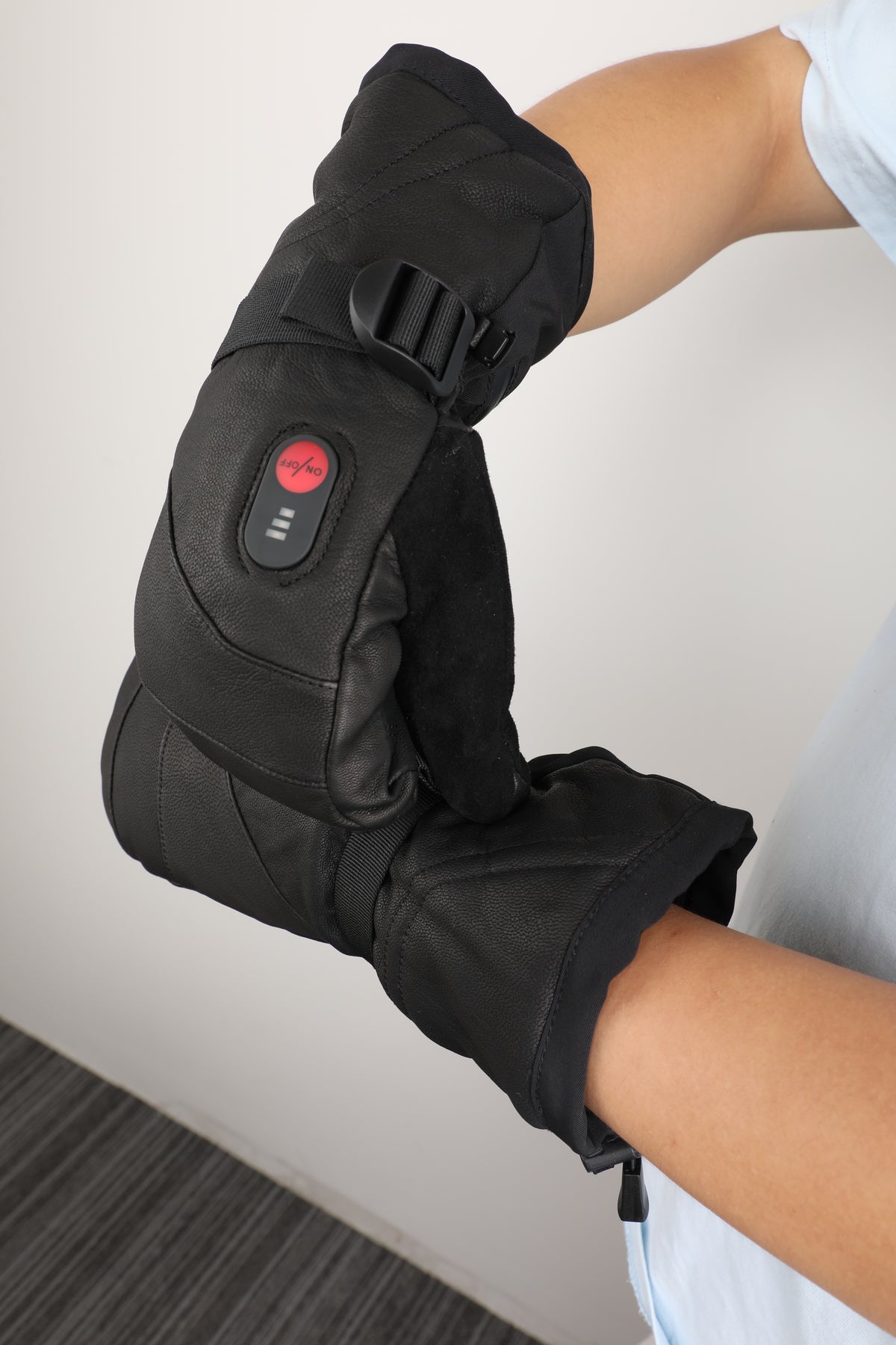 A person wearing a black heated knee sleeve with an integrated control panel featuring a red power button, shown against a neutral indoor background.